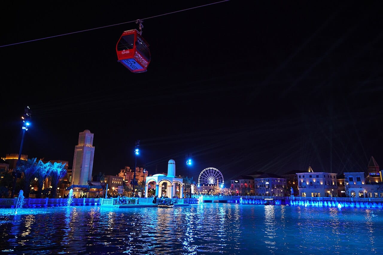 A cable car over the lit lake and pavilions of Boulevard World during Riyadh Season