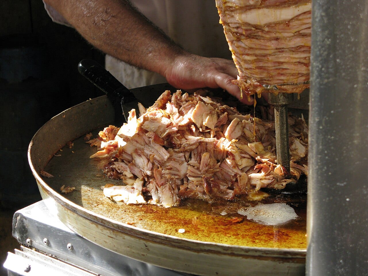 A cook shaving chicken shawarma from a vertical spit onto a hot tray